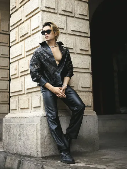 An edgy, high-fashion look for a male model, featuring leather and a confident pose against a stone pillar.