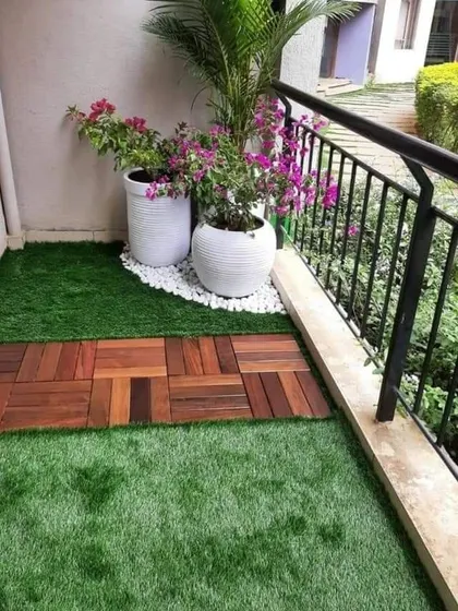 Adding color and life to a small balcony space. This setup features a combination of artificial grass and wooden deck tiles, with flowering bougainvillea and palms in classic white planters.