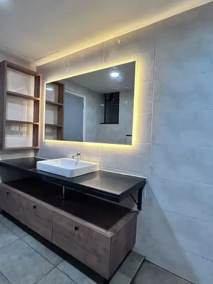 A modern bathroom vanity unit with a large, LED-backlit mirror. The design includes a floating wooden cabinet and open shelving, providing ample storage with a clean, contemporary look.