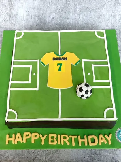 A football field cake for a birthday celebration. The design features a jersey with the birthday boy's name and number, along with a football, all on an edible pitch.