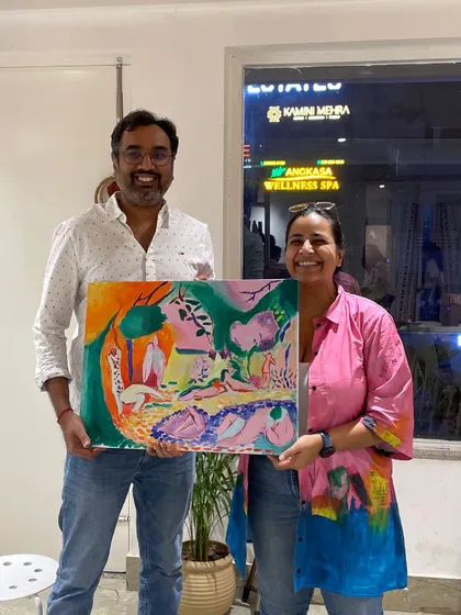 A couple holds up their vibrant and colorful paintings, one a large abstract piece and the other a smaller landscape.
