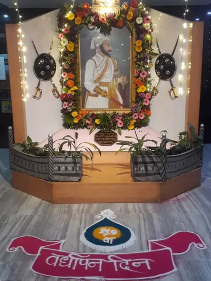 A respectful and beautiful setup for a cultural event, celebrating 'Vardhapan Din'. The decor includes a floral frame for the portrait, traditional shields, and a colorful rangoli.