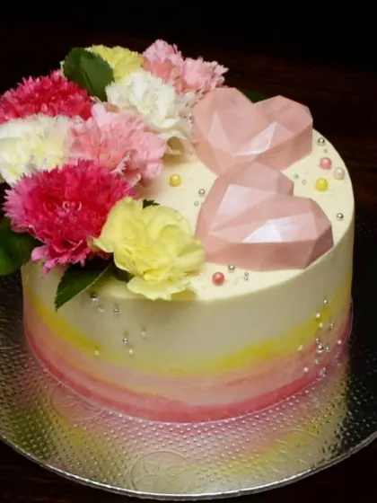 This pretty anniversary cake features a soft watercolor blend of pink and yellow buttercream. It's decorated with fresh carnations and two pink geometric chocolate hearts for a modern and romantic look.