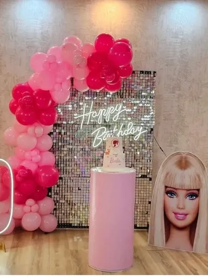 A chic Barbie birthday setup with a pink balloon arch against a silver shimmer wall, a '9' neon number, and a Barbie head cutout.