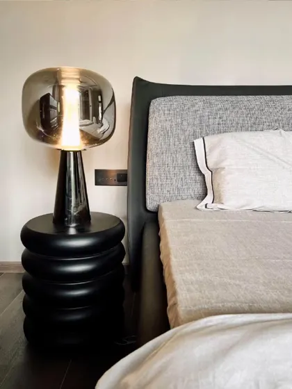 A close-up of a bedside setup, showcasing a sculptural black side table and a modern lamp with a smoked glass shade. The design focuses on the beauty of simple forms and the contrast between the grey fabric headboard and the dark wood floor.