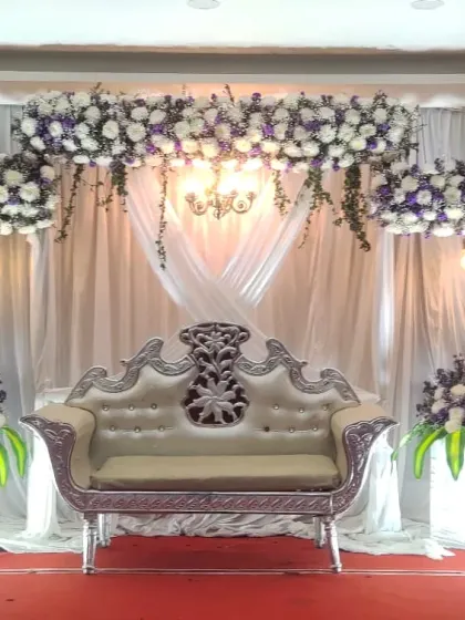 A classic baby shower stage with white and purple floral arrangements, warm lighting, and an ornate silver-and-white sofa.