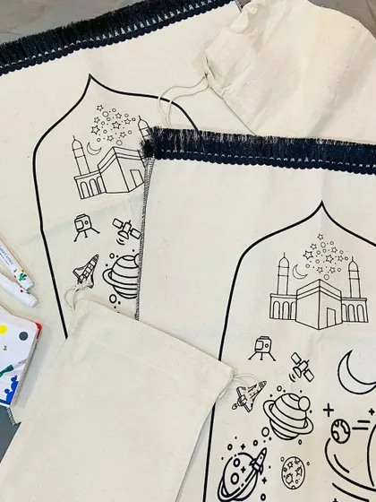 A closer look at the "Colour, Create, Pray" prayer mat for kids. The design features the Kaaba, planets, and stars, ready to be colored in with the included fabric markers, making prayer time a creative and engaging experience.