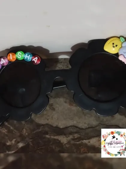 A close-up of the personalized kids' sunglasses. These flower-shaped frames are decorated with the name 'Naisha' in letter beads and other cute charms.
