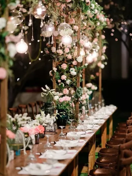 An intimate and romantic long-table dining setup for a reception. The table is decorated with an overhead structure of flowers and hanging light bulbs.