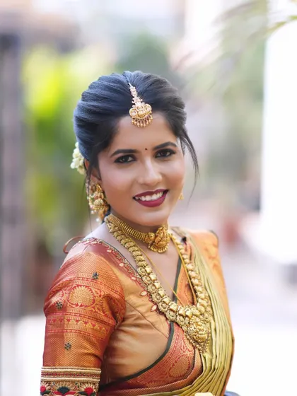 A radiant bridal portrait. The hairstyle is a traditional South Indian bun adorned with flowers, complementing her intricate gold jewelry and the rich colors of her muhurtham saree.