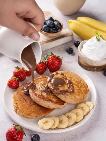 An action shot of chocolate sauce being poured over a stack of fluffy pancakes, creating a moment of pure indulgence.