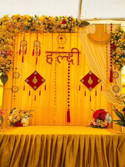 This Haldi backdrop combines traditional yellow drapes with unique decorative hangings and the auspicious 'Haldi' sign in Hindi script.