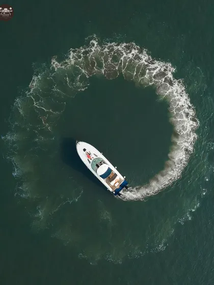 A stunning top-down aerial view of a white yacht creating a perfect circle with its wake in the dark green ocean. This creative drone shot turns a pre-wedding moment into a piece of art.