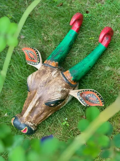 A wooden bull head, hand-painted with colorful details, seen through foliage. These pieces are inspired by temple festival traditions and add an authentic, festive touch to any space.