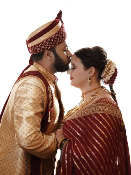 A tender moment captured as the groom kisses the bride's forehead. The clean white background puts all the focus on the couple's affection and their ornate wedding clothes.