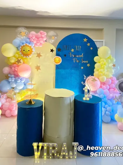 A "First Trip Around The Sun" space-themed birthday party. This cute setup features astronaut and rocket cutouts, a sun and stars backdrop, and a pastel-colored balloon garland.