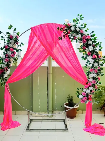 For smaller functions or outdoor events, I can provide a simple and elegant metal arch. This one is decorated with bright pink drapes, artificial flower arrangements, and fairy lights, serving as a beautiful and budget-friendly photo point.