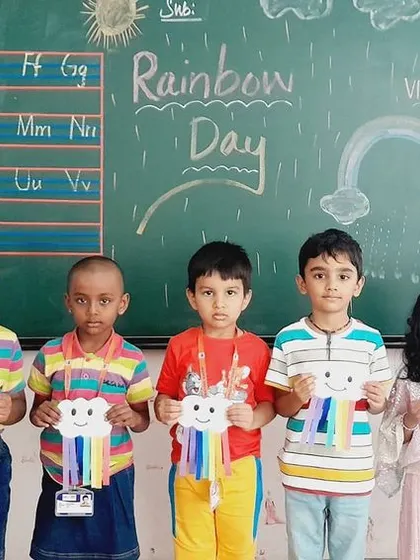 Students stand in front of a blackboard decorated for Rainbow Day, holding their artwork.