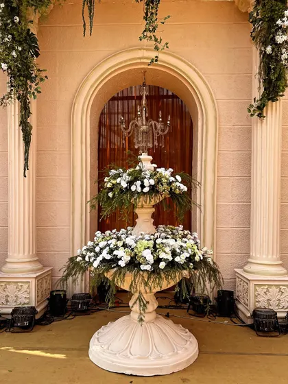 A grand floral fountain serves as a centerpiece in this Roman-themed decor. The two-tiered structure overflows with white flowers and green foliage, flanked by classical pillars.
