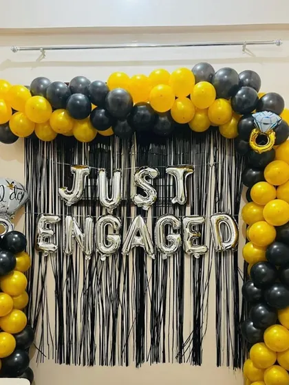 A bold and stylish 'Just Engaged' photo booth. The backdrop features a black tinsel curtain framed by a black and yellow balloon arch, with silver foil balloons announcing the occasion.