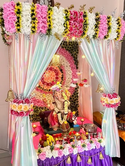 A vibrant and colorful Ganpati mandap featuring a multi-layered floral backdrop in concentric circles. The iridescent fabric and hanging bells add a playful and festive touch to the celebration.