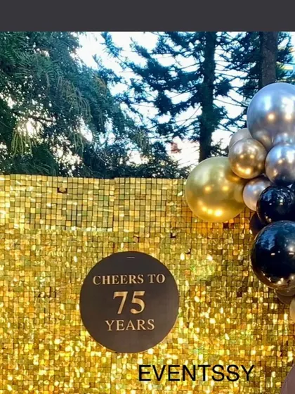 A glamorous "Cheers to 75 Years" photo backdrop. The shimmering gold sequin wall provides a perfect background for photos, complemented by a classic black, gold, and silver balloon garland.