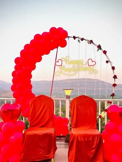 A romantic anniversary setup on a terrace with a view, featuring a red balloon arch and covered chairs.