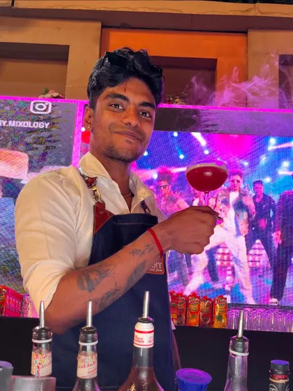 A bartender proudly presents a smoky, glowing red cocktail, with a vibrant LED screen in the background adding to the party atmosphere.