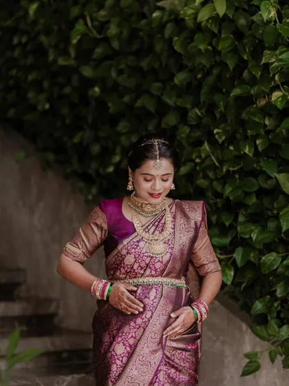 A full-length portrait showcasing the elegance of a traditional maternity look. The rich purple saree and intricate gold jewellery are beautifully complemented by the natural green foliage in the background.