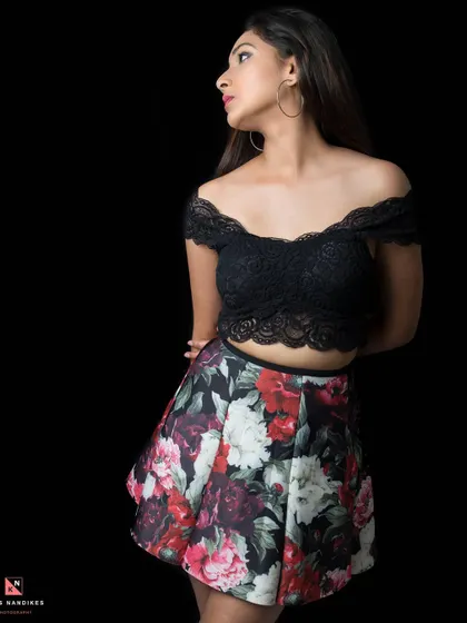 A low-key studio portrait where the model wears a black lace top and a floral skirt. The lighting is focused on her, making her stand out against the pure black background.