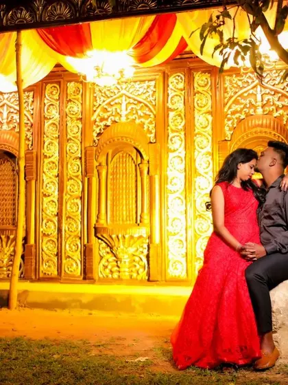 A couple sits together in front of a grand, golden, illuminated backdrop at night. The bride's red dress and the opulent setting create a regal feel.