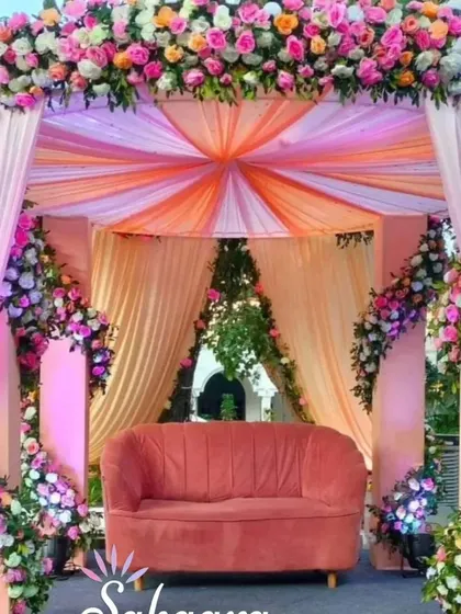 A beautiful outdoor wedding mandap draped in pastel peach and pink fabric. The structure is covered in lush floral arrangements, creating a soft and romantic setting for a daytime wedding.