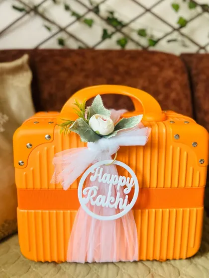 The exterior of a bright orange mini suitcase hamper, decorated with a "Happy Rakhi" acrylic tag and a delicate white bow.