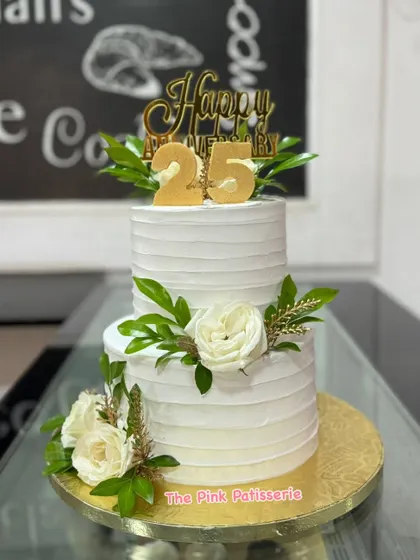 A classic and elegant two-tiered cake for a 25th anniversary. The clean, white textured buttercream is beautifully decorated with fresh white roses, greenery, and a gold "25" topper.