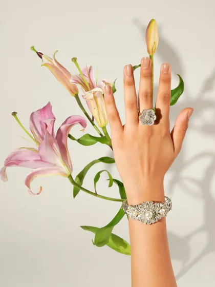A model's hand, adorned with a floral-inspired ring and bracelet, is elegantly posed with fresh lilies. This image blends the beauty of nature with the intricate craftsmanship of fine jewellery.