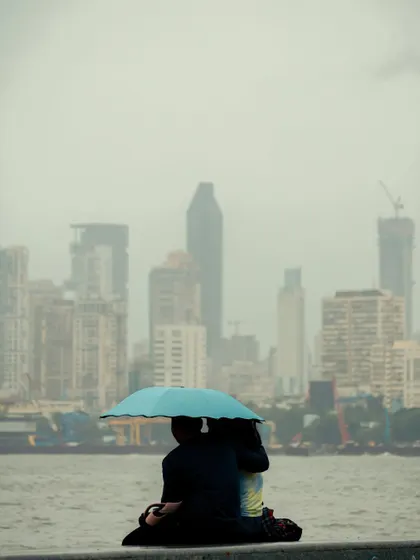 A couple sits by the sea under a bright blue umbrella, with the city skyline hazy in the distance. The image evokes a sense of shared intimacy and escape from the city.