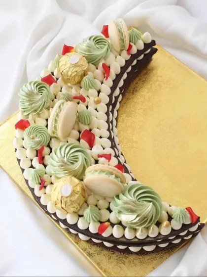 A beautiful crescent moon-shaped cake, perfect for Eid or Ramadan celebrations. This chocolate cake is decorated with green and white buttercream swirls, macarons, and Ferrero Rocher.