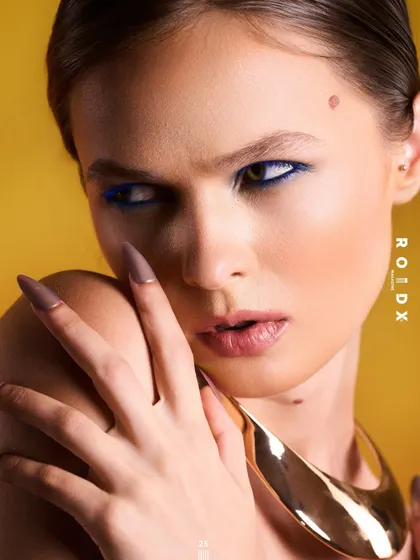 This cover for Roidx Magazine is a powerful beauty and accessory shot. The sharp lighting highlights the model's features, the bold blue eyeliner, and the smooth, sculptural gold neckpiece, creating a look of modern femininity.