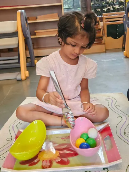 Transferring objects with a large spoon is a classic practical life activity that builds fine motor control and concentration. This child is learning precision and coordination, skills that are essential for writing and other detailed tasks.
