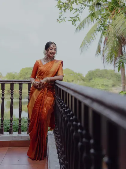 The bride, dressed in a beautiful orange Kanjeevaram saree, smiles as she poses on a balcony overlooking a serene backwater setting.
