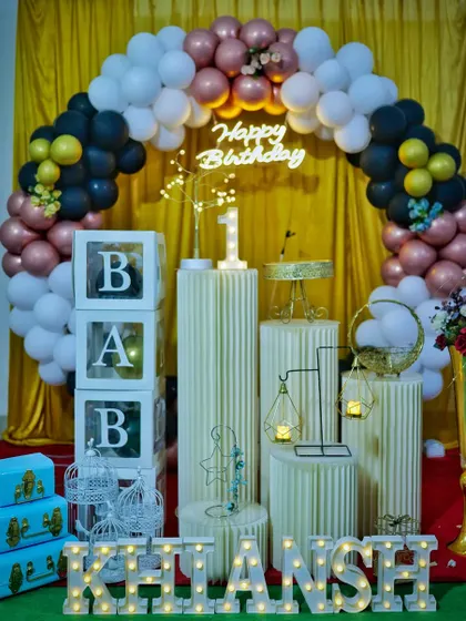 A classic first birthday setup with a black, white, and rose gold balloon arch against a gold curtain backdrop.
