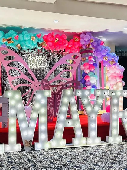 A low-angle shot of a colorful butterfly-themed birthday stage. The large marquee letters for "AMAYRA" are the centerpiece, surrounded by butterfly wings and multi-colored balloons.