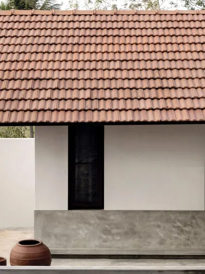 The simple, clean lines of the exterior, featuring a traditional Mangalore tile roof, white walls, and a cement plaster base. The design includes a gravel path for effective rainwater drainage.