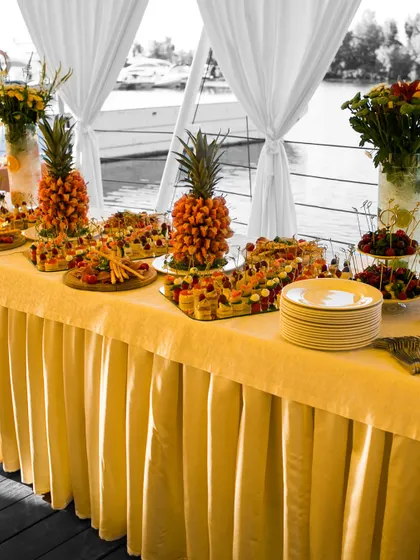 A vibrant fruit and appetizer buffet set up for an event by the water. The display features pineapple fruit towers and a colorful array of canapés, creating a fresh and inviting spread for guests.