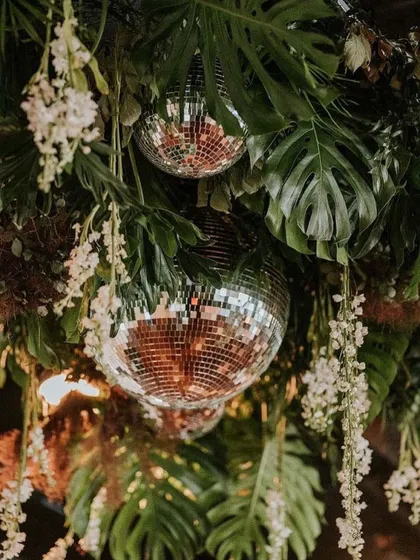 A creative ceiling decoration where disco balls are nestled among tropical leaves and hanging white flowers, perfect for a jungle-glam themed party.