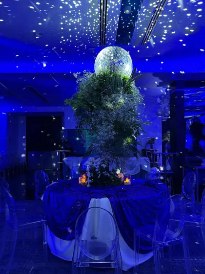 A beautifully set table at the blue-themed birthday party. A large centerpiece with a disco ball and baby's breath sits on a table draped in velvet, under a ceiling of twinkling lights that look like a galaxy.
