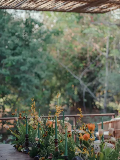A close-up of a rustic, autumnal tablescape on our wooden deck. The arrangement features wildflowers, dried elements, and green candles in dark wooden holders.