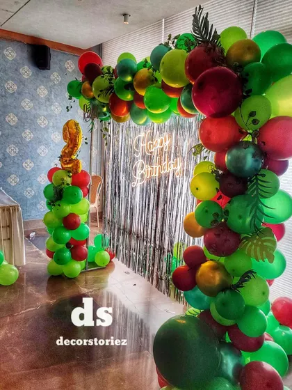 A stylish jungle-themed balloon arch with a neon 'Happy Birthday' sign. The mix of different green and red tones, along with faux tropical leaves, creates a modern and chic look.