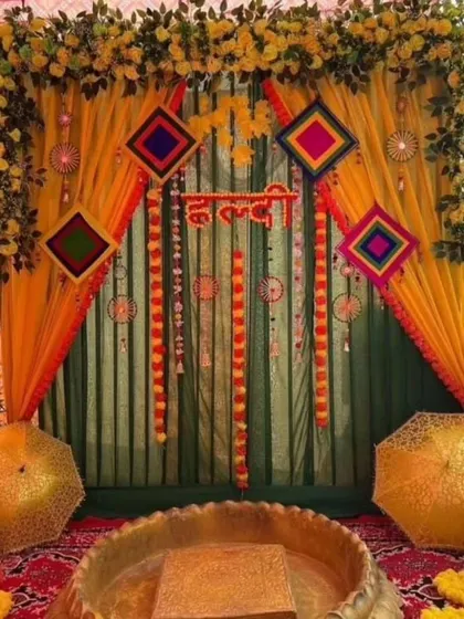 A traditional Haldi ceremony setup with a green and yellow draped backdrop, decorated with colorful hangings and umbrellas. The arrangement includes a large brass 'urli' for the rituals.