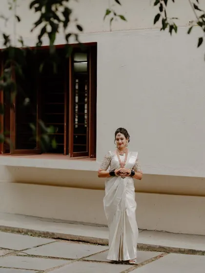 A wide shot of a bride in a white saree standing before a building, framed by delicate leaves. This composition adds an artistic touch to the bridal portrait.